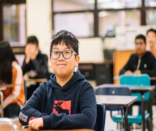 Batang Bata na may salamin na nakaupo sa isang silid-aralan na nakangiti